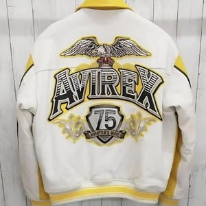 K&ouml;nnte beinhalten: Eine wei&szlig;-gelbe Collegejacke mit einer gro&szlig;en Stickerei auf der R&uuml;ckseite. Das Motiv zeigt einen Adler, den Schriftzug AVIREX, ein Wappen mit der Zahl 75 und den Text AVIATOR'S KING. Die Jacke hat einen gelben Kragen und gelbe S&auml;ume.