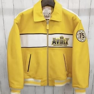 K&ouml;nnte beinhalten: Eine leuchtend gelbe Jacke mit einem wei&szlig;en horizontalen Bruststreifen, einem durchgehenden Frontrei&szlig;verschluss und einem Umlegekragen. Auf der Brust befindet sich das gestickte Avirex Aviator's King Logo, am linken &Auml;rmel ein runder Aufn&auml;her mit der Zahl 75.