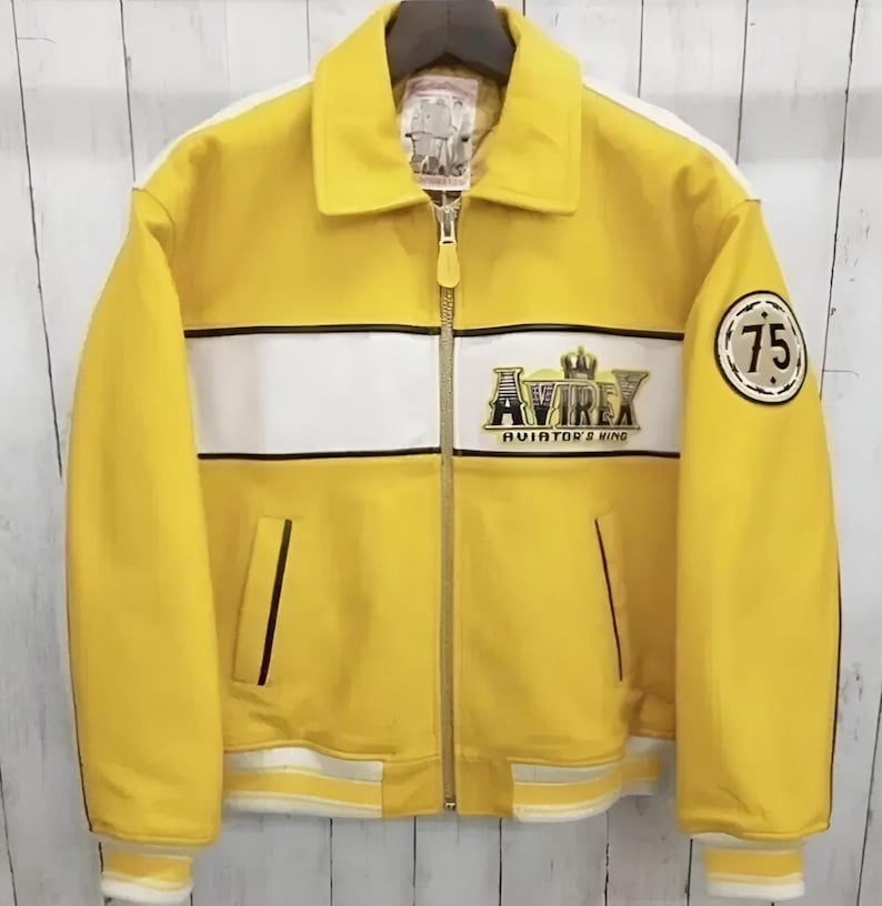 K&ouml;nnte beinhalten: Eine leuchtend gelbe Jacke mit einem wei&szlig;en horizontalen Bruststreifen, einem durchgehenden Frontrei&szlig;verschluss und einem Umlegekragen. Auf der Brust befindet sich das gestickte Avirex Aviator's King Logo, am linken &Auml;rmel ein runder Aufn&auml;her mit der Zahl 75.