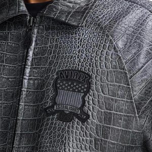 May include: A charcoal gray jacket with a textured crocodile skin pattern. The jacket features a front zipper closure and a black embroidered patch on the chest that reads AVIREX ESTABLISHED 1975 with a shield graphic containing stars and stripes.