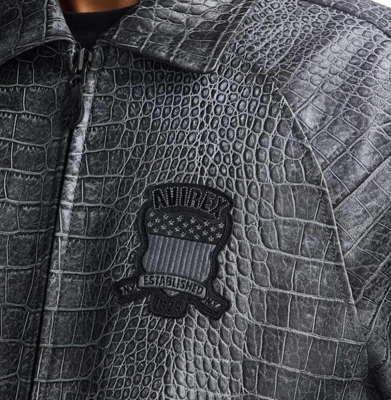 May include: A charcoal gray jacket with a textured crocodile skin pattern. The jacket features a front zipper closure and a black embroidered patch on the chest that reads AVIREX ESTABLISHED 1975 with a shield graphic containing stars and stripes.