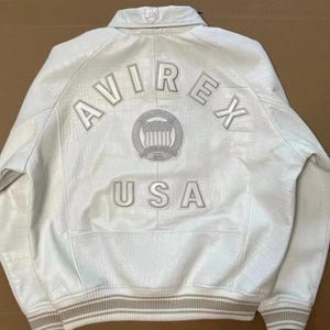May include: A white leather jacket with a textured crocodile-skin pattern. The back features the embroidered text AVIREX USA in white letters, along with a circular emblem containing a shield and stars. It has a ribbed collar and cuffs.