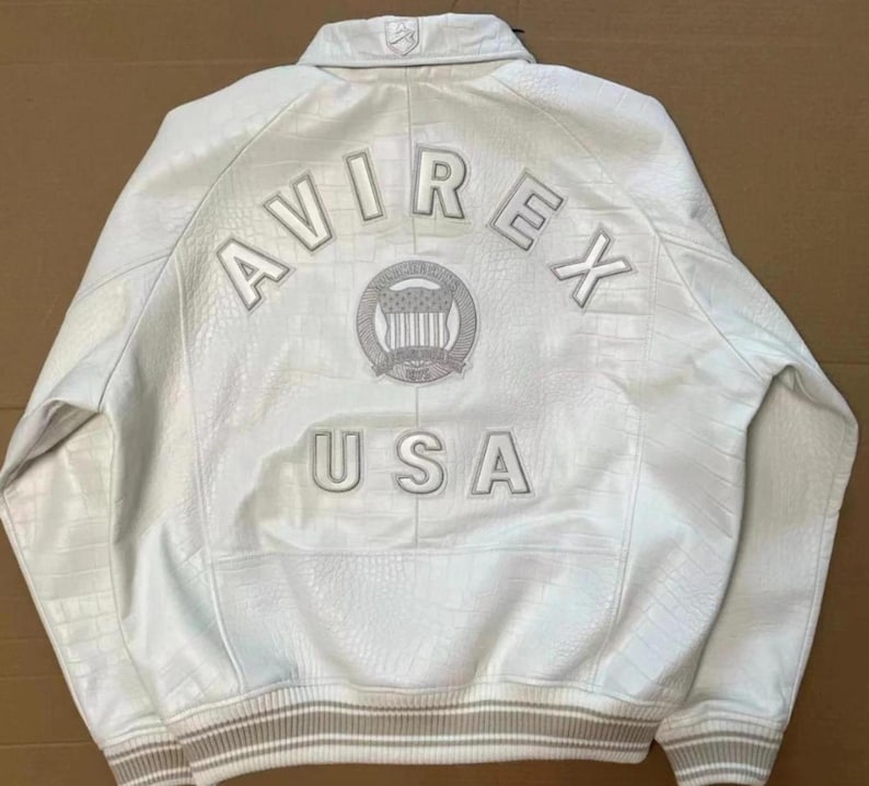 May include: A white leather jacket with a textured crocodile-skin pattern. The back features the embroidered text AVIREX USA in white letters, along with a circular emblem containing a shield and stars. It has a ribbed collar and cuffs.
