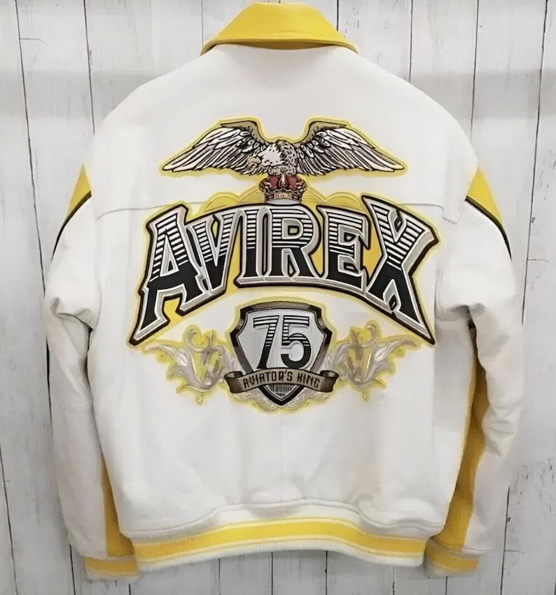 K&ouml;nnte beinhalten: Eine wei&szlig;-gelbe Collegejacke mit einer gro&szlig;en Stickerei auf der R&uuml;ckseite. Das Motiv zeigt einen Adler, den Schriftzug AVIREX, ein Wappen mit der Zahl 75 und den Text AVIATOR'S KING. Die Jacke hat einen gelben Kragen und gelbe S&auml;ume.