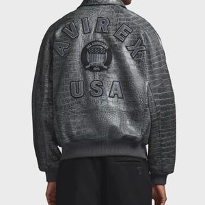 May include: A charcoal gray jacket with a textured crocodile skin pattern. The back features the text AVIREX in an arched layout, the word USA below it, and a circular patch in the center with the text ALL AMERICANS ESTABLISHED 1975.