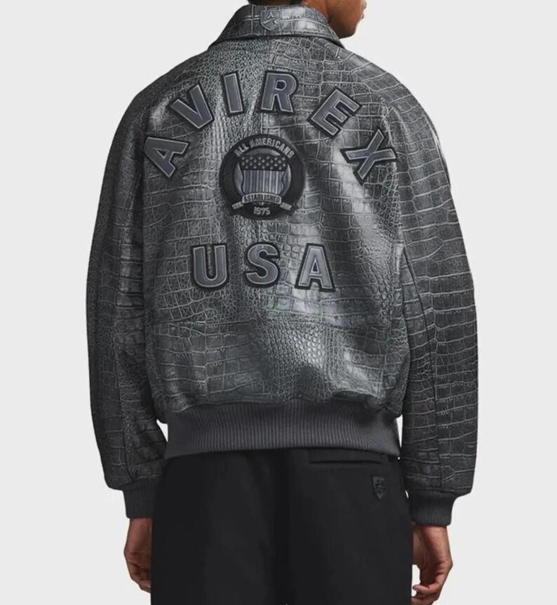 May include: A charcoal gray jacket with a textured crocodile skin pattern. The back features the text AVIREX in an arched layout, the word USA below it, and a circular patch in the center with the text ALL AMERICANS ESTABLISHED 1975.