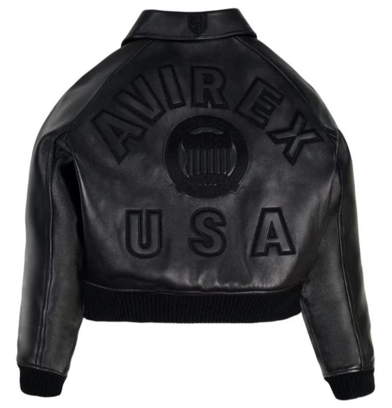 May include: A black leather jacket featuring a classic collar and ribbed cuffs and hem. The back displays the embossed text AVIREX USA in large capital letters, centered around a circular emblem with a shield design, all in a matching black tone.