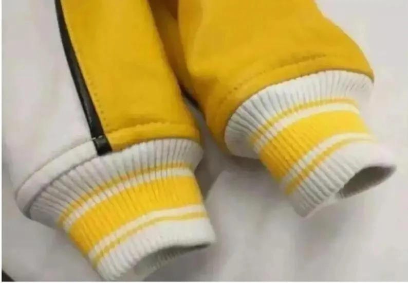 K&ouml;nnte beinhalten: Ein &Auml;rmeldetail einer Jacke mit einem leuchtend gelben und einem wei&szlig;en Bereich, die durch eine d&uuml;nne schwarze vertikale Linie getrennt sind. Der &Auml;rmel endet in einem gerippten Strickb&uuml;ndchen in Wei&szlig; mit zwei horizontalen gelben Streifen.