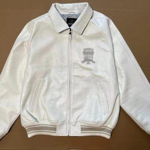 May include: A white jacket with a textured finish, a full-length front zipper, and a pointed collar. The chest features an embroidered emblem with the text AVIREX 1975. The cuffs and hem are finished with a beige and white striped ribbed knit trim.