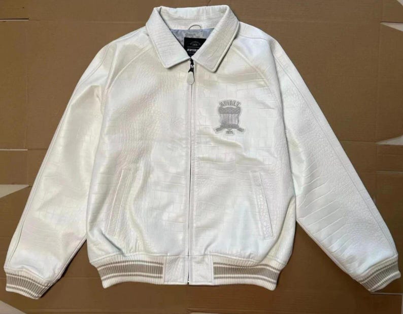 May include: A white jacket with a textured finish, a full-length front zipper, and a pointed collar. The chest features an embroidered emblem with the text AVIREX 1975. The cuffs and hem are finished with a beige and white striped ribbed knit trim.