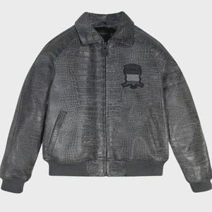 May include: A charcoal gray bomber jacket featuring a textured crocodile skin pattern throughout. The jacket includes a front zipper closure, a standard collar, ribbed cuffs and hem, and a black embroidered patch on the left chest area.