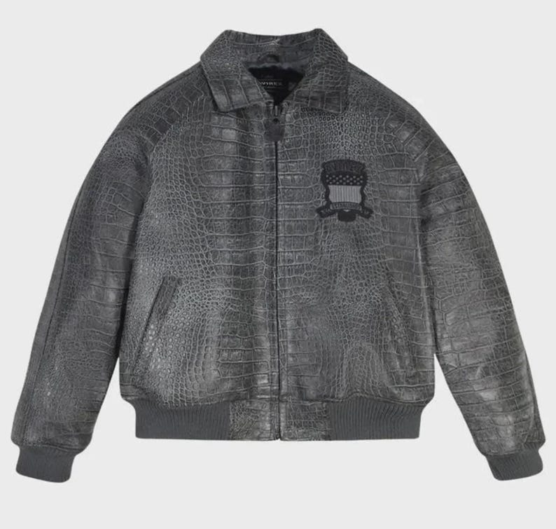 May include: A charcoal gray bomber jacket featuring a textured crocodile skin pattern throughout. The jacket includes a front zipper closure, a standard collar, ribbed cuffs and hem, and a black embroidered patch on the left chest area.