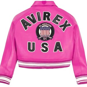 Op de afbeelding: Een felroze varsity-jack met de zwarte tekst AVIREX in een boog boven een ronde patch met een Amerikaanse vlag en de tekst ALL AMERICANS ESTABLISHED 1975. De zwarte tekst USA staat onder de patch, met wit en roze gestreepte geribbelde boorden.
