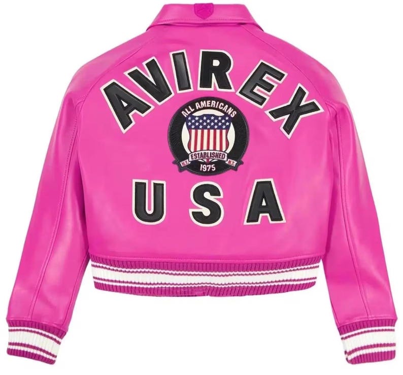 Op de afbeelding: Een felroze varsity-jack met de zwarte tekst AVIREX in een boog boven een ronde patch met een Amerikaanse vlag en de tekst ALL AMERICANS ESTABLISHED 1975. De zwarte tekst USA staat onder de patch, met wit en roze gestreepte geribbelde boorden.