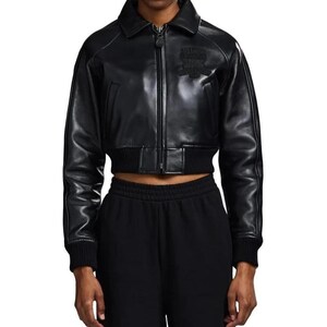 May include: A black cropped jacket with a smooth finish, featuring a pointed collar, a central front zipper, and ribbed knit cuffs and hem. The jacket includes a tonal embroidered emblem on the upper left chest and long sleeves with seam detailing.