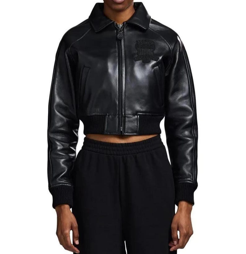 May include: A black cropped jacket with a smooth finish, featuring a pointed collar, a central front zipper, and ribbed knit cuffs and hem. The jacket includes a tonal embroidered emblem on the upper left chest and long sleeves with seam detailing.