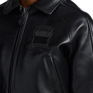 May include: A black leather jacket featuring a classic collar, a full-length front zipper with a branded pull tab, and a black embroidered patch on the chest. The patch displays the text AVIREX, ESTABLISHED, and 1975, along with a shield graphic.