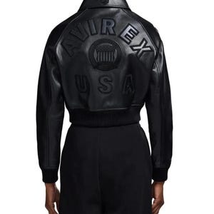 May include: A black leather jacket featuring a classic collar and ribbed cuffs. The back displays embossed lettering that reads AVIREX USA, along with a circular emblem containing a shield graphic. The jacket has a structured, cropped fit.