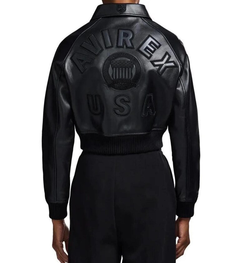 May include: A black leather jacket featuring a classic collar and ribbed cuffs. The back displays embossed lettering that reads AVIREX USA, along with a circular emblem containing a shield graphic. The jacket has a structured, cropped fit.