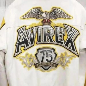 K&ouml;nnte beinhalten: Eine wei&szlig;e Jacke mit einem gro&szlig;en gestickten R&uuml;ckenaufn&auml;her, der ein gelb-schwarzes AVIREX-Logo, einen gekr&ouml;nten Wei&szlig;kopfseeadler und ein Schild mit der Zahl 75 &uuml;ber dem Schriftzug AVIATOR'S KING sowie gelbe und graue Details zeigt.