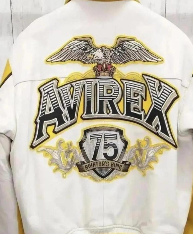 K&ouml;nnte beinhalten: Eine wei&szlig;e Jacke mit einem gro&szlig;en gestickten R&uuml;ckenaufn&auml;her, der ein gelb-schwarzes AVIREX-Logo, einen gekr&ouml;nten Wei&szlig;kopfseeadler und ein Schild mit der Zahl 75 &uuml;ber dem Schriftzug AVIATOR'S KING sowie gelbe und graue Details zeigt.