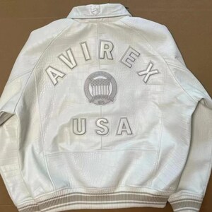 May include: A white leather jacket with a textured crocodile-like pattern. The back features the text AVIREX in an arc above a circular emblem containing a shield and stars, with the text USA below. It has a ribbed collar and hem.