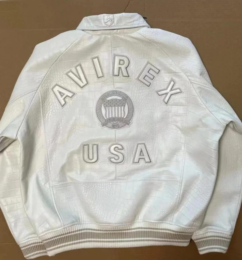 May include: A white leather jacket with a textured crocodile-like pattern. The back features the text AVIREX in an arc above a circular emblem containing a shield and stars, with the text USA below. It has a ribbed collar and hem.