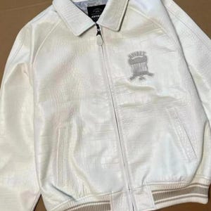 May include: A white jacket with a textured crocodile-embossed pattern, featuring a full-length front zipper, a pointed collar, and ribbed cuffs. An embroidered Avirex shield logo is on the left chest, and the interior label reads Avirex 1975.
