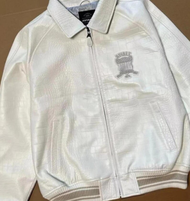 May include: A white jacket with a textured crocodile-embossed pattern, featuring a full-length front zipper, a pointed collar, and ribbed cuffs. An embroidered Avirex shield logo is on the left chest, and the interior label reads Avirex 1975.