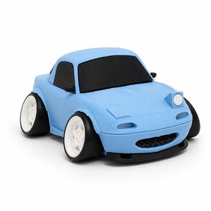 May include: A light blue toy car with a stylised, compact design. It features black windows, a black front bumper, and wide white wheels with black tyres. The car has pop-up headlights, a rounded body shape, and a small black grille on the front.