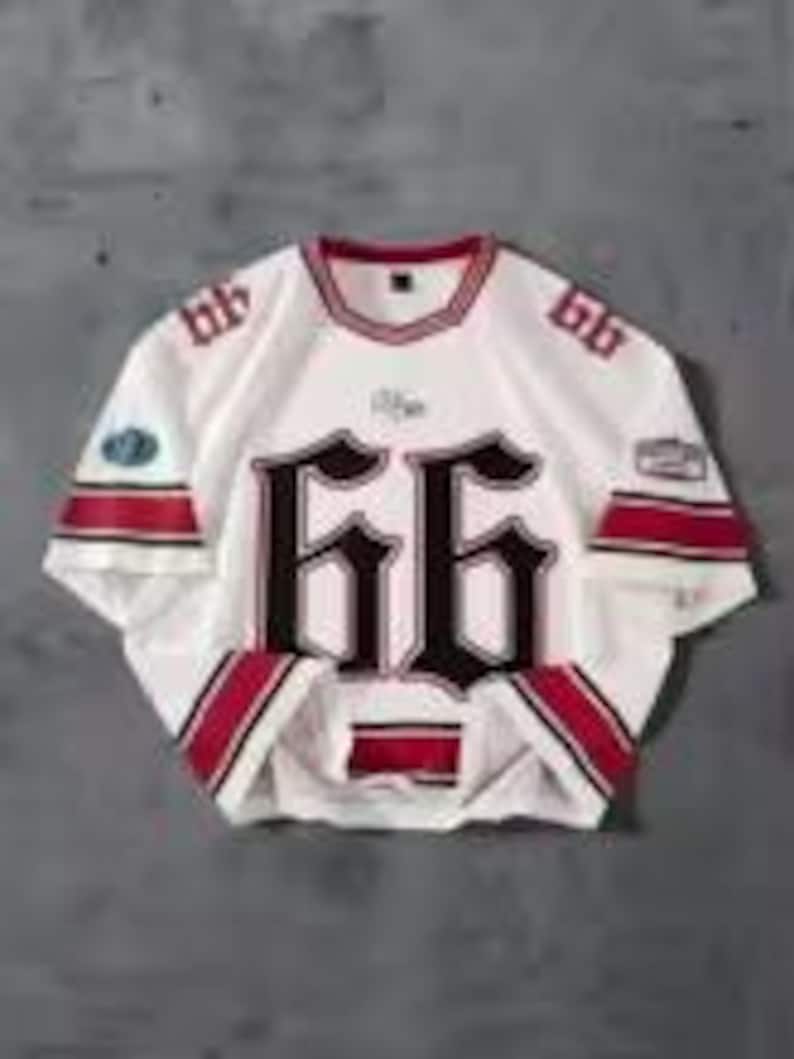 May include: White hockey jersey with red and black accents. The number "66" is prominently displayed on the front and sleeves in a gothic font. The jersey has a round neckline and a patch on the left sleeve.