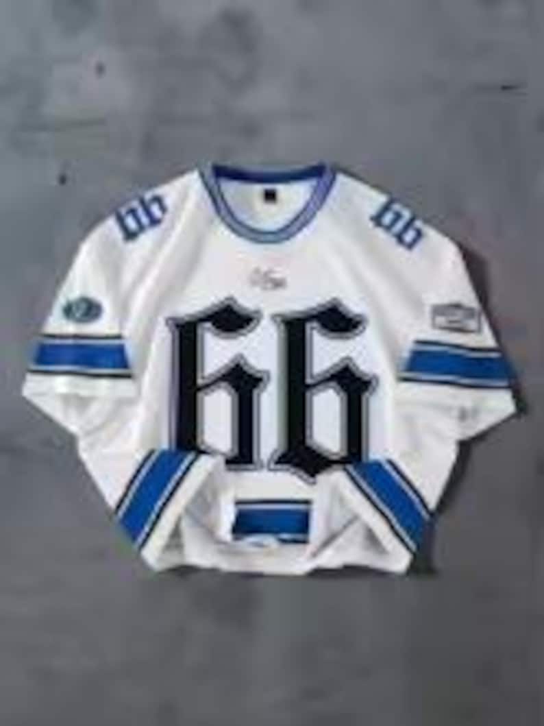 May include: White jersey with blue and black accents, featuring the number "66" in large, gothic-style font on the front. The jersey has blue stripes on the sleeves and collar, with patches on the sleeves. The jersey is displayed on a gray surface.