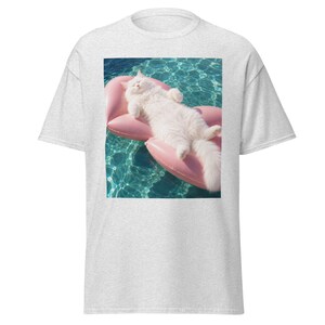 May include: A light heather grey t-shirt featuring a rectangular graphic print of a fluffy white cat lying on a pink heart-shaped inflatable pool float. The cat is positioned in the centre of the image, floating on clear blue swimming pool water.
