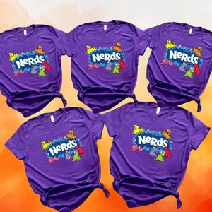 May include: Five purple short-sleeve t-shirts arranged in a group. Each shirt features the white text "NERds" centered on the chest, surrounded by a colorful graphic of small, cartoonish characters in yellow, orange, pink, purple, and green.