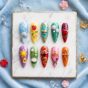 May include: A set of ten long, almond-shaped nail designs. Each nail features a different design, including a carriage, rose, genie lamp, seashells, apple, sun, frog, fan, crown, and bow and arrow. The nails are set on a white marble surface.