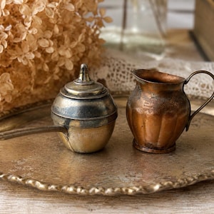 Peut inclure: Un récipient rond en laiton avec couvercle et long manche, accompagné d'un petit pot à lait en cuivre au corps cannelé et anse incurvée, posés sur un plateau circulaire en laiton à la finition texturée, patinée et au bord festonné.