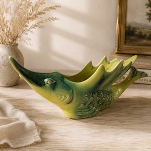 Peut inclure: Un bol en céramique en forme de poisson stylisé avec un museau pointu et une nageoire caudale. La pièce présente un vernis dégradé passant d'un vert forêt profond au niveau de la tête à un jaune citron vif sur le corps, avec des écailles moulées.