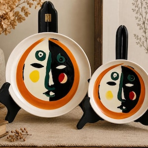 May include: Two decorative ceramic plates with a face design. The plates feature a white background with an orange border and a face design in black, green, yellow, and red. The plates are displayed on black stands.
