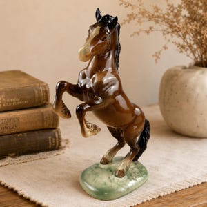May include: A glossy ceramic horse figurine in a rearing pose, featuring a rich brown coat with a white blaze on its face and black mane and tail. The horse stands on a textured, light green oval base, showcasing detailed muscle definition.