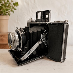 Puede incluir: Una cámara plegable vintage Zeiss Ikon con cuerpo negro de textura de cuero, fuelle negro y un conjunto de lente de metal plateado. La cámara cuenta con un visor montado en la parte superior, un brazo de soporte cromado y un barril de lente.