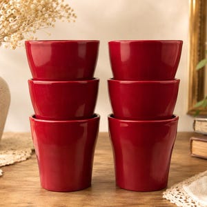May include: Six glossy, red ceramic bowls are stacked in two columns of three. The bowls have a slightly flared shape. The bowls are on a wooden surface, with a vase and framed artwork in the background.
