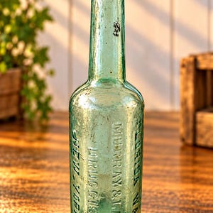 May include: A vintage light green glass bottle with a long neck and a rounded body. Embossed text on the front reads FLORIDA WATER, MURRAY & LANMAN, DRUGGISTS, NEW YORK. The glass surface shows signs of age with minor imperfections and texture.