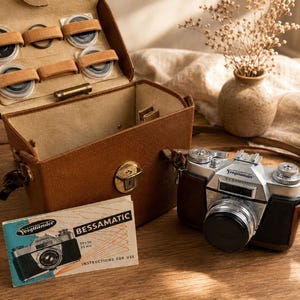 May include: A vintage Voigtländer Bessamatic camera with a brown leather case, a matching brown leather camera bag with internal lens storage, and an instruction manual. The camera features a silver and black body with a lens cap attached.