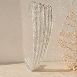 May include: A clear glass cuff bracelet featuring a textured, rippled surface with vertical ridges. The accessory has an open-ended design, allowing it to be slipped on the wrist. The glass has a translucent quality with a rough, bubbly finish.