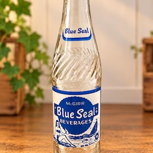 May include: A clear glass bottle with a narrow neck and a textured spiral pattern. The label features the text Blue Seal and McGirr Blue Seal Beverages in blue ink, accompanied by a graphic illustration of a person and a sailboat on the water.