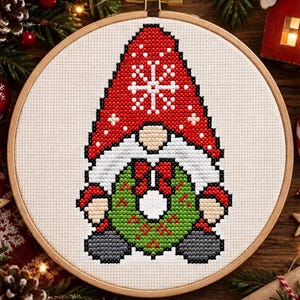 May include: A cross-stitch embroidery in a round wooden hoop featuring a gnome with a tall red hat decorated with white snowflakes. The gnome has a white beard, tan skin, and is holding a green wreath with red berries and a red bow.