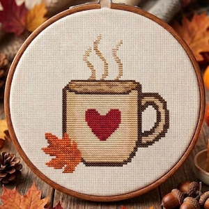 May include: A circular wooden embroidery hoop containing a cross-stitch design of a tan mug with a dark red heart on the front. The mug features steam rising from the top and an orange maple leaf positioned at the base of the mug on the left side.