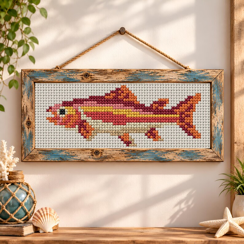 May include: A rectangular cross-stitch artwork featuring a fish with scales in shades of red, orange, yellow, and cream. The piece is encased in a weathered, distressed wooden frame with blue and brown tones, suspended by a thick jute rope.