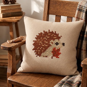 May include: A square beige throw pillow featuring a cross-stitch design of a hedgehog. The hedgehog is rendered in shades of brown, tan, and orange, with a small dark brown nose and eye. The pillow fabric has a visible grid-like texture.