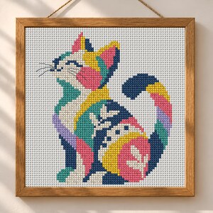 May include: A square cross-stitch embroidery of a cat in a wooden frame. The cat is composed of geometric color blocks in navy blue, bright yellow, pink, teal, and lavender, featuring white leaf-like patterns on its body and a white background.